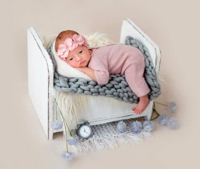 Charming newborn in floral diadem lying awake on tiny bed