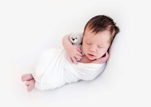 Adorable newborn in white blanket holding toy in tiny hand