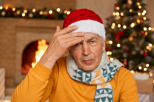 Tired unhappy annoyed old man being sick on Christmas eve, suffering from headache, keeping hand on forehead, wearing yellow shirt and santa hat, looks at camera with upset expression.
