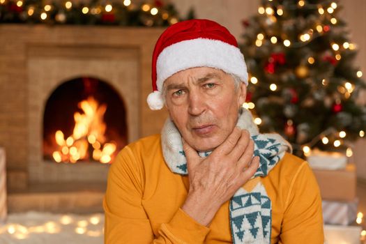 Tired or sick old man in Santa Claus hat, touching neck feeling pain and fever, getting ill on Christmas holiday, posing on background with blurred fireplace, xmas tree and gift boxes.