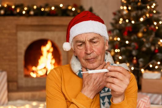 Unhealthy senior man in santa hat sitting near fireplace wrapped in scarf, holding thermometer in hands, has fever, posing in festive room with Christmas decoration and fireplace.