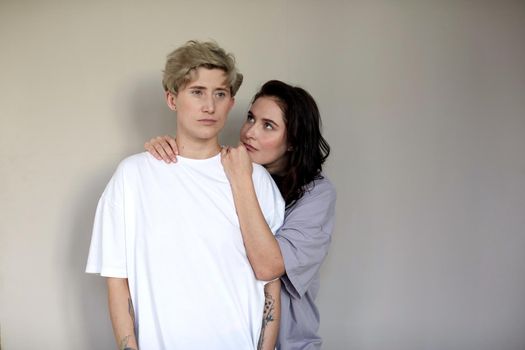 Young brunette embracing and looking at androgynous woman with short hair against gray wall