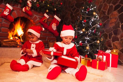 Little girl and boy are sitting near fireplace and christmas tree with gift boxes. Brother and sister in santa suits