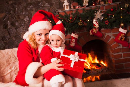 Mother and son are sitting near fireplace and christmas tree. Family look into a gift box.