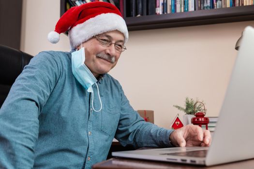 Christmas online congratulation. Senior man in santa claus hat talks using laptop for video call friends and childrens. The room is festively decorated. Christmas during coronavirus.