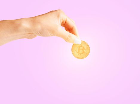 Woman’s hand holding glowing gold bitcoin on a pink background, copy-space for text.