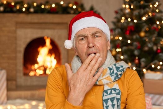 Sleepy man in yellow sweater, scarf and Christmas hat yawning, covering mouth with hand on background of fireplace and fir tree, Happy New Year, celebration holiday at home.