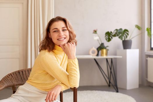Content female with charming smile leaning on hand and looking at camera while sitting on wicker chair at home