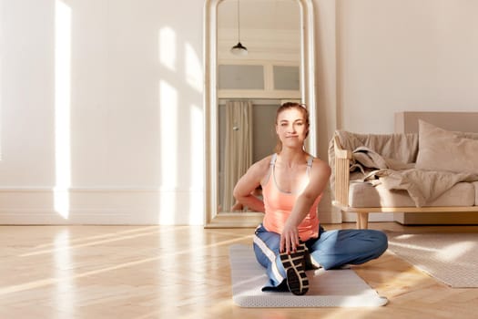 Smiling female athlete in sportswear sitting on mat in living room and stretching legs while warming up before workout at home and looking at camera