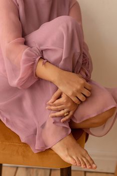 High angle of anonymous barefoot female in pink translucent dress sitting on chair and embracing knee