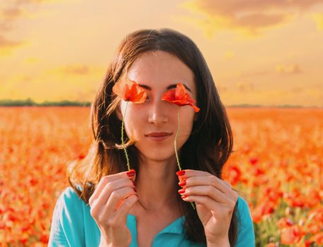 Beautiful young woman holding red poppies in front of her eyes and standing in flower meadow at sunset.