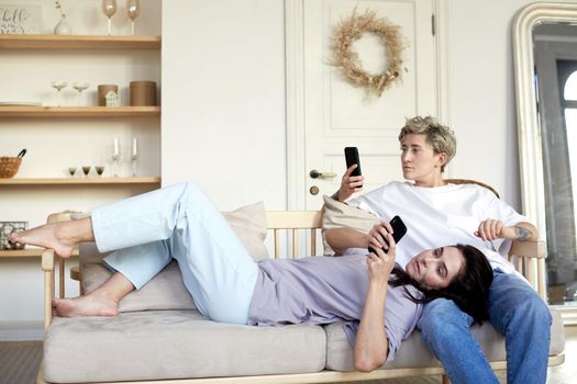 Young women resting on couch and browsing mobile phones together in cozy living room in weekend at home