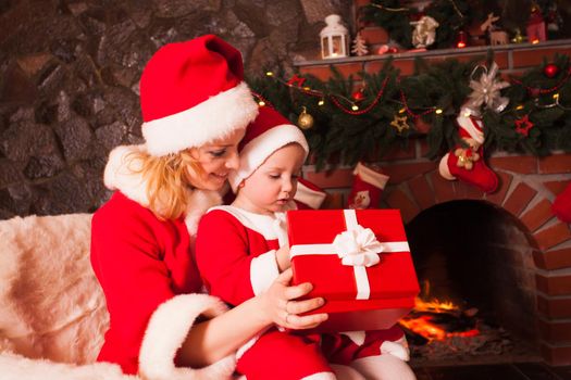 Mother and son are sitting near fireplace and christmas tree. Family look into a gift box.