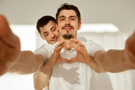 Loving man showing heart while embracing boyfriend from behind taking selfie in light apartment room