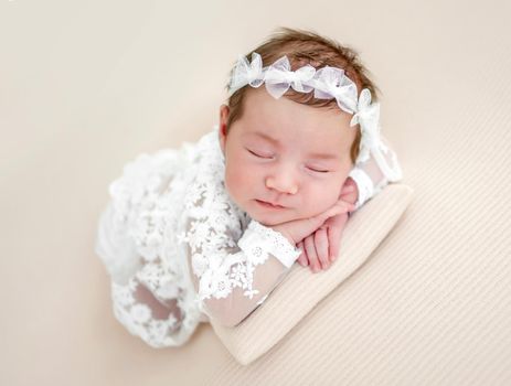 Closeup portrait of adorable newborn baby girl wearing beautiful white dress and wreath lying on her tummy and sleeping in studio. Cute infant child napping holding hands under cheeks