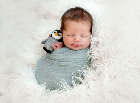 Newborn baby boy sleeping swaddled in fabric on white fur and holding knitted toy in tiny hands. Infant kid studio portrait with decoration