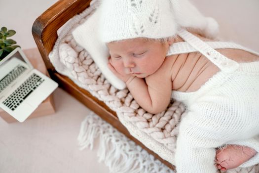 Newborn baby boy sleeping in tiny bed wearing knitted white costume and hat. Infant kid studio portrait with tiny toy laptop