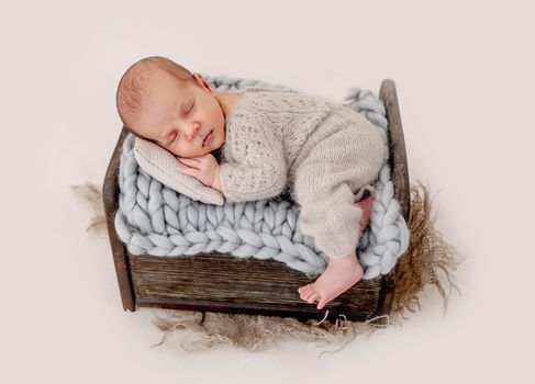 Adorable newborn baby boy sleeping on knitted blanket in tiny wooden bed and holding his hands under cheek. Infant child napping during studio photoshoot