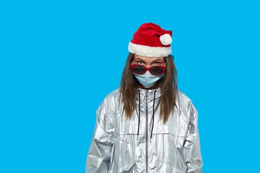 Serious female wearing Santa hat and protective mask and red sunglasses standing on blue background in studio and looking at camera. Lady looks over the glasses