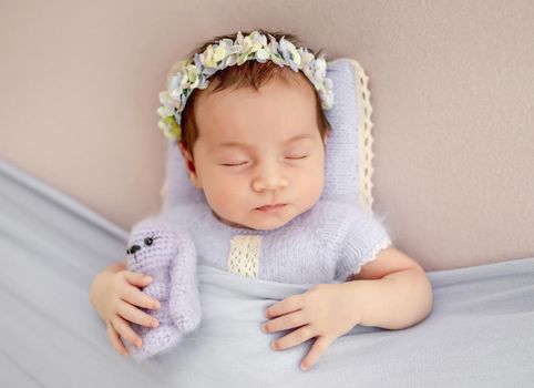 Beautiful little newborn baby girl wearing knitted costume and wreath with flowers holding toy in her tiny hands and sleeping under blanket in studio. Cute infant child napping