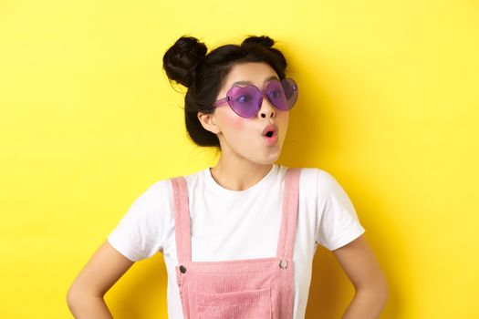 Summer and fashion concept. Close-up of surprised asian teen girl in heart-shaped sunglasses looking right side with excited face, standing on yellow background.