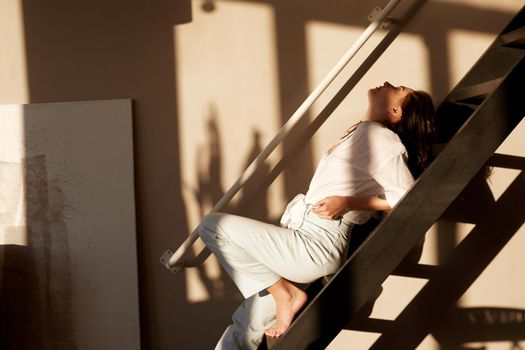 Side view of sensual young female with dark hair in casual clothes leaning on stairs with closed eyes during rest in art studio