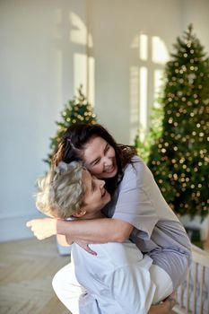 Cheerful young women laughing and embracing each other while celebrating Christmas in cozy room at home