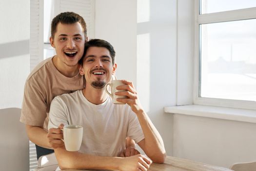 Cheerful young men with mugs of hot drinks smiling and looking at camera while sitting at table in morning at home