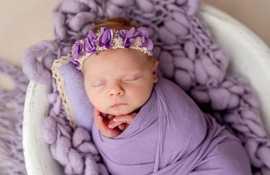 Newborn wearing floral rim sleeping wrapped in violet blanket in cradle