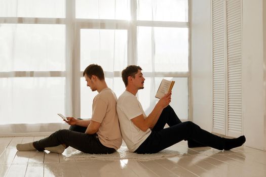 Side view of young boyfriends using smartphone and reading book while sitting back to back against window on weekend day at home