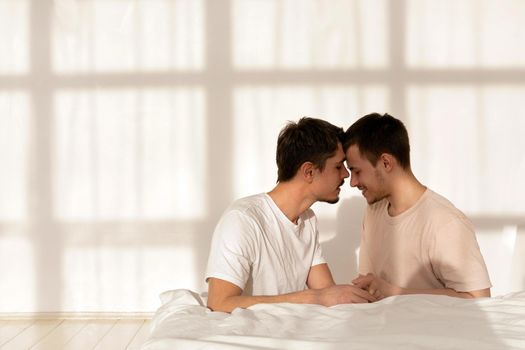Cute gay couple touch their foreheads with closed eyes against the background of a white wall in a bedroom on a sunny morning