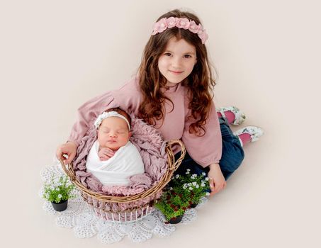 Beautiful little girl sitting with her swaddled beautiful newborn baby sister sleeping in the basket with fur on light pink background. Adorable kid napping and her sibling staying close to her
