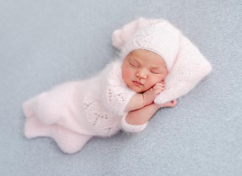 Cute newborn baby girl in knitted hat and costume sleeping on grey background and holding her hands under cheek. Adorable infant child napping during studio photoshoot