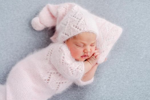 Cute newborn baby girl in knitted hat and costume sleeping on grey background and holding her hands under cheek. Adorable infant child napping during studio photoshoot. Little kid closeup portrait