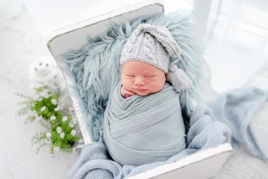 Portrait of newborn baby boy swaddled in light blue fabric and wearing knitted hat sweet sleeping on white small designed bed. Adorable infant child napping during studio photoshoot
