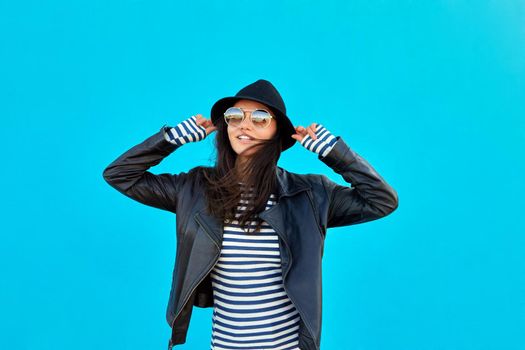 Delighted trendy female in black leather jacket and sunglasses touching hat blue background