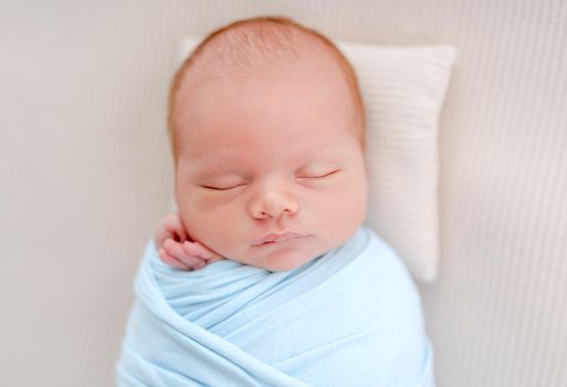 Portrait of newborn baby boy swaddled in light blue fabric sleeping on white background and holding tiny hands under his cheeks. Adorable infant child napping during studio photoshoot