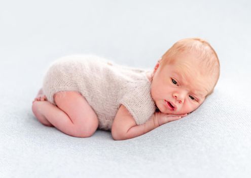 Newborn baby boy wearing knitted beige costume lying on his tummy and holding tiny hands under his cheeks. Adorable infant child resting and looking at the camera
