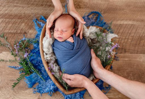 Hands of father and mother holding adorable newborn baby boy swaddled in blue fabric during sleeping. Cute infant kid napping in fur in wooden heart bed during studio photoshoot