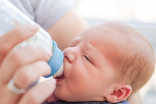 Newborn baby boy lying in mother hands and eating milk from bottle. Matherinity care about sleeping infant kid. Closeup portrait of little child