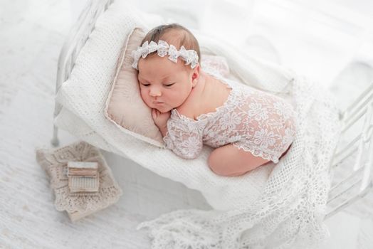 Beautiful newborn baby girl wearing lace costume ana wreath lying on her tummy on stylized white bed with pillow during studio photoshoot. Cute portrait of infant child in white room from above