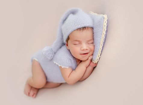 Adorable newborn baby girl wearing knitted costume and hat sleeping holding hands under her cheeks on pillow in studio. Cute infant child napping and smiling