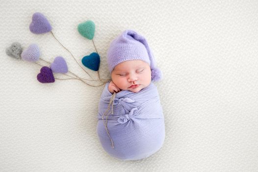 Newborn baby girl swaddled in light blue fabric sleeping and holding in her hands knitted colorful hearts. Little cute infant child napping in beautiful composition. Adorable kid wearing hat resting