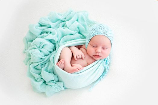 Adorable newborn baby girl wearing knitted hat lying on her back in the cocoon on white background and sleeping. Sweet infant child napping during studio photoshoot with decoration