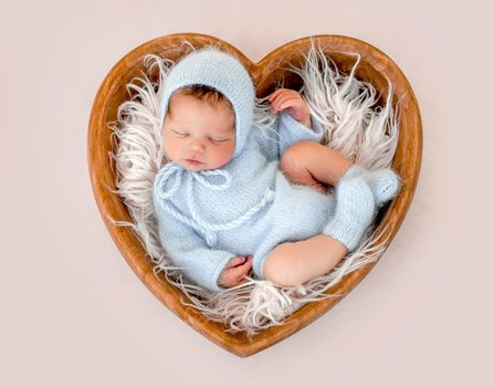 Newborn baby boy sleeping wearing knitted costume and hat in tiny heart shape bed. Infant kid studio portrait with fur
