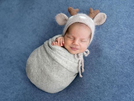 Adorable newborn baby boy swaddled in fabric and wearing cute knitted hat sleeping during studio photoshoot. Infant child napping