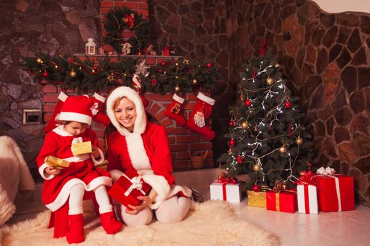 Mother and daughter are sitting near fireplace and christmas tree with gift boxes.