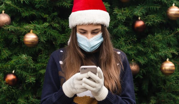 Christmas online greetings. Close up Portrait of woman wearing a santa claus hat and medical mask with emotion. Against the background of a Christmas tree. Coronavirus pandemic