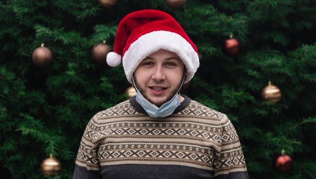 Wearing a mask incorrectly. Close up Portrait of man wearing a santa claus hat, xmas sweater and medical mask with emotion. Against the background of a Christmas tree. Coronavirus pandemic