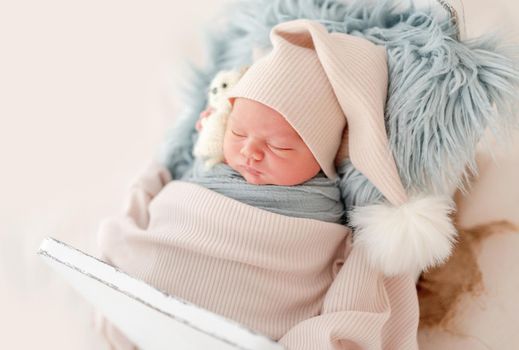 Newborn baby boy wearing knitted hat sleeping on white small designed bed. under blanket Adorable infant child napping during studio photoshoot